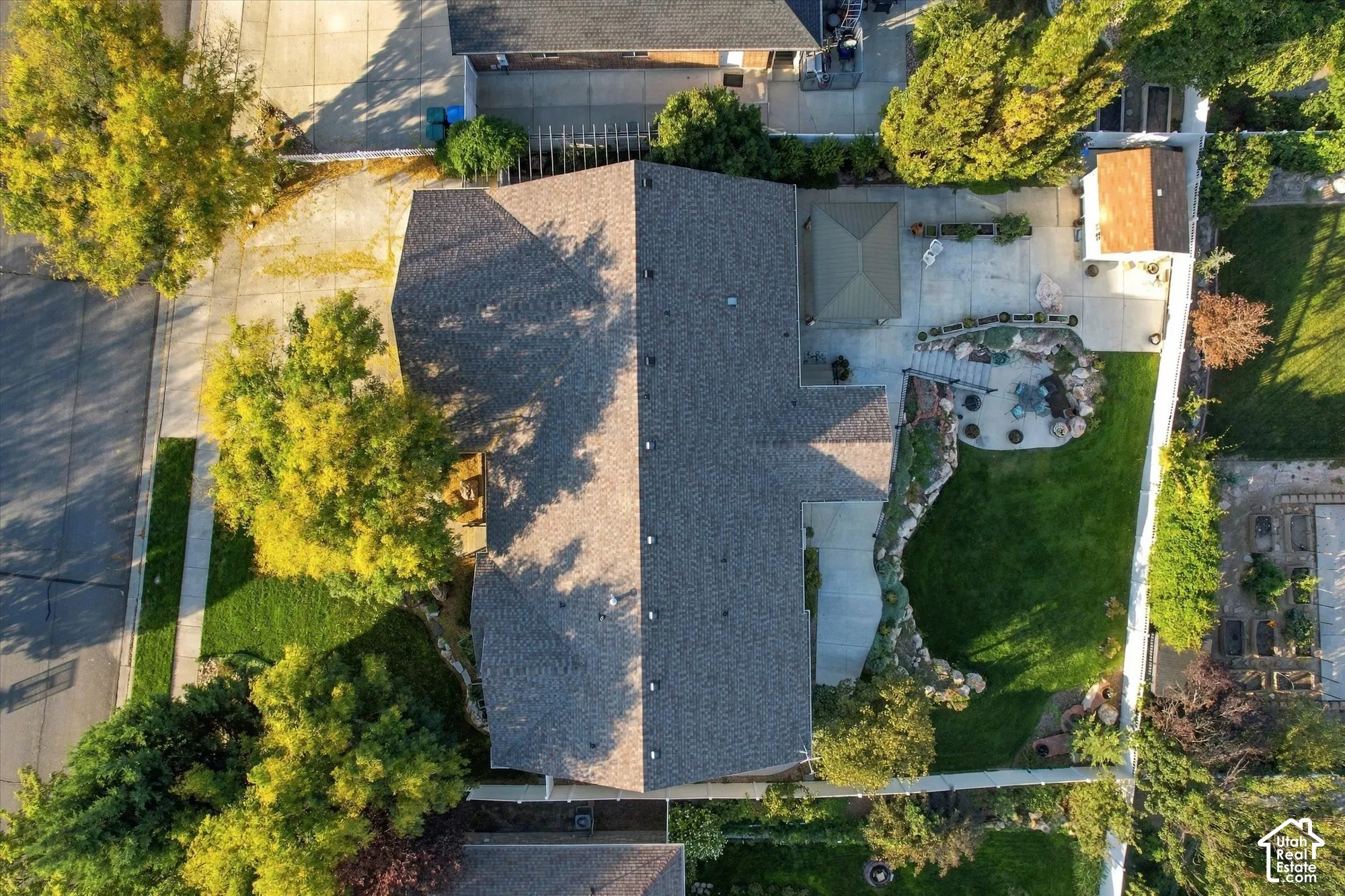 Aerial view showcasing a substantial corner-lot property with a charcoal-gray asphalt shingle roof, white exterior siding, and mature landscaping featuring established trees and manicured lawn areas