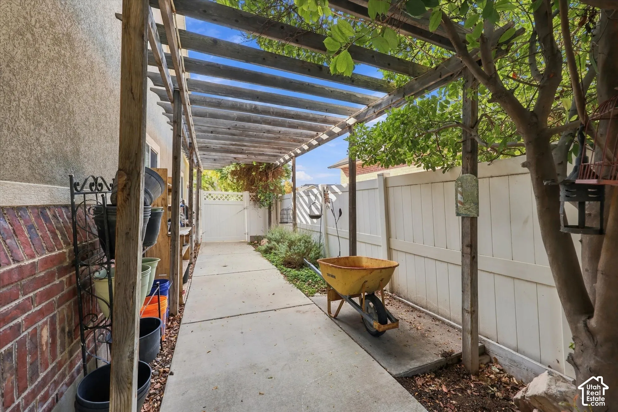 Wood pergola structure extending over a concrete pathway, bordered by a brick wall and a white panel fence