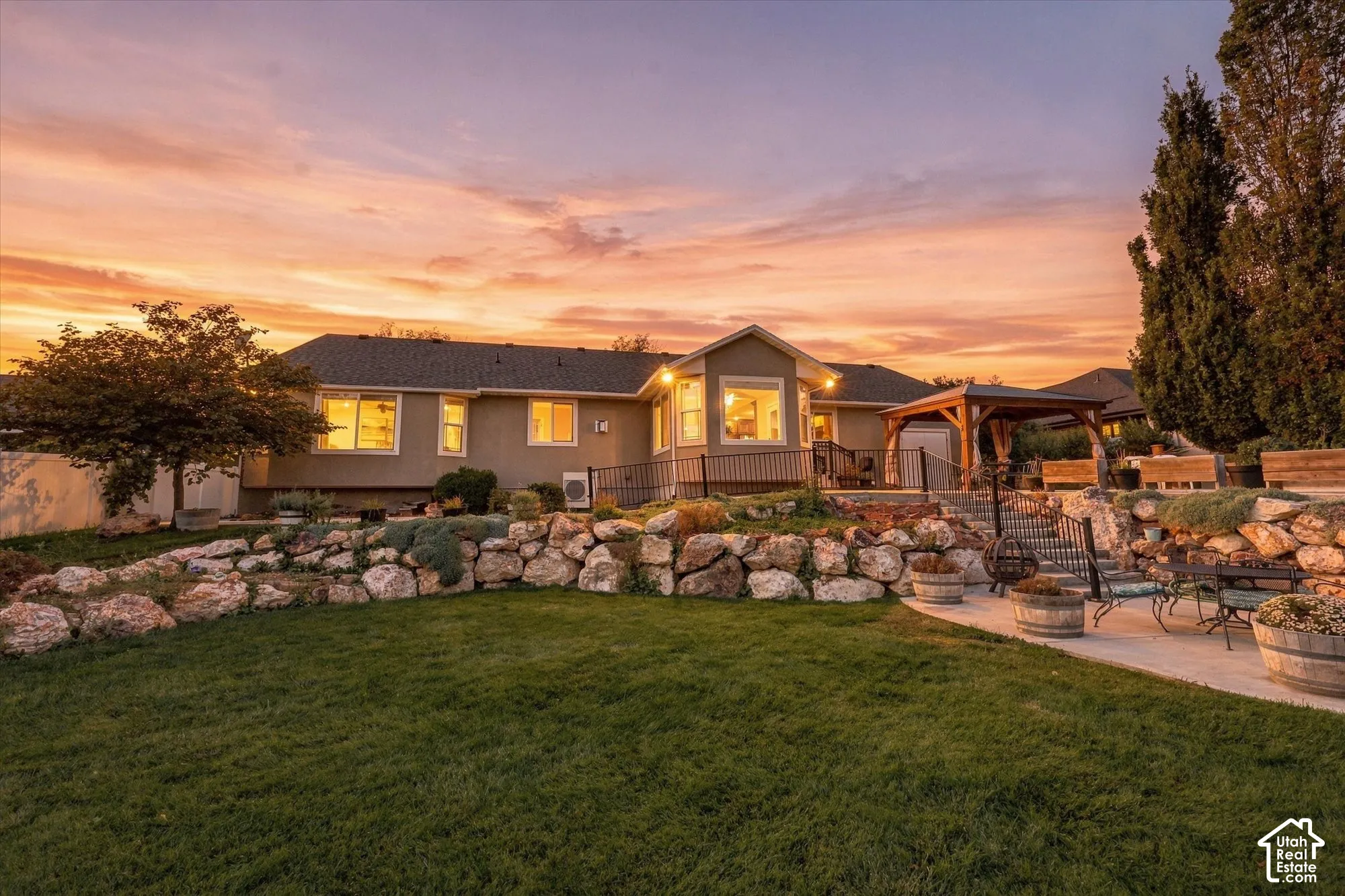 Twilight elegance defines this residence, featuring a well-lit exterior with multiple illuminated windows, a manicured lawn, mature landscaping with specimen trees, decorative boulder border accents, and an expansive patio area ideal for outdoor entertain