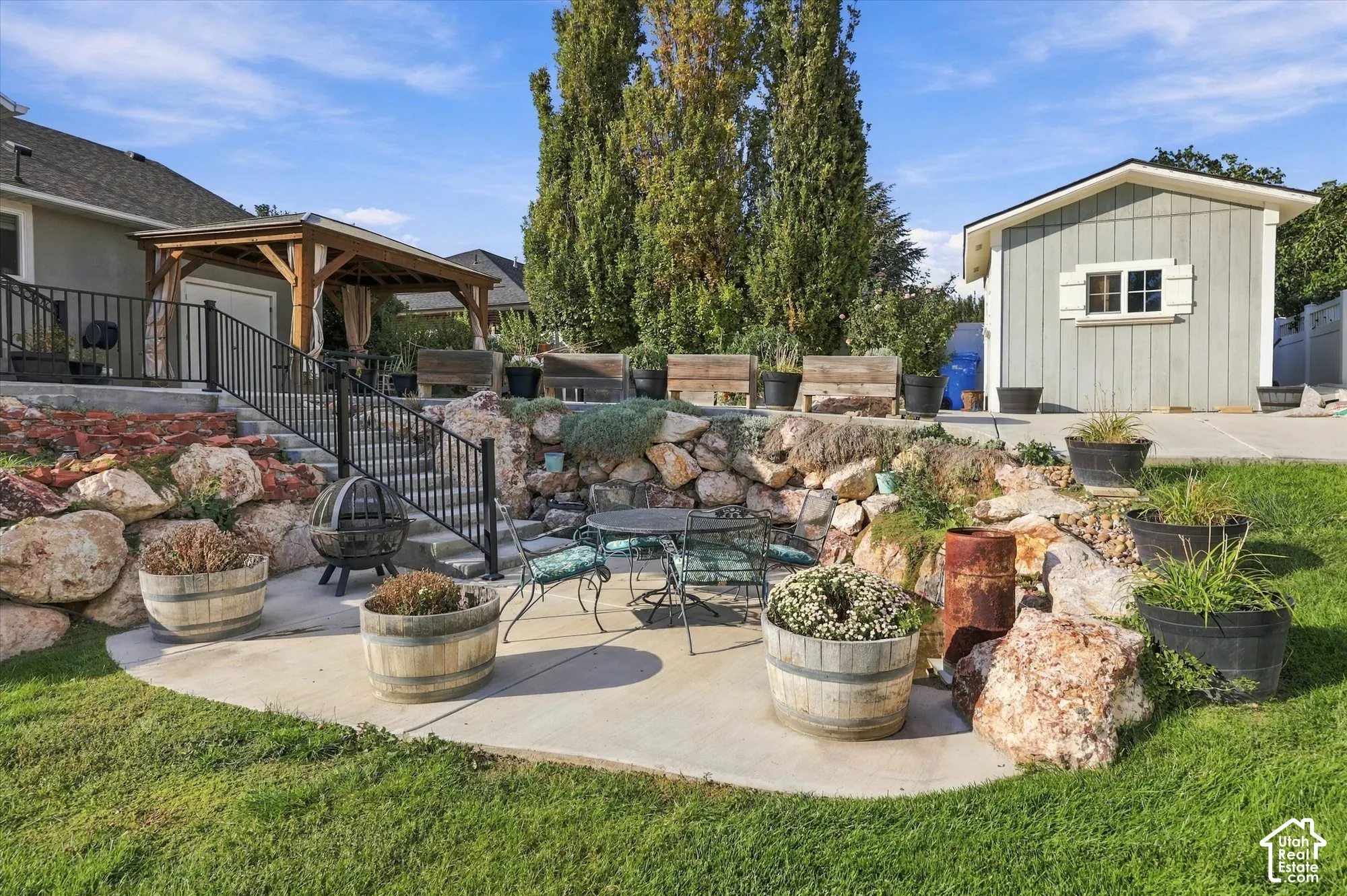 Expansive backyard oasis featuring a concrete patio with integrated seating area, decorative boulder landscaping, mature trees including a tall Italian cypress, multiple large planters with seasonal plantings, wooden pergola structure attached to the home