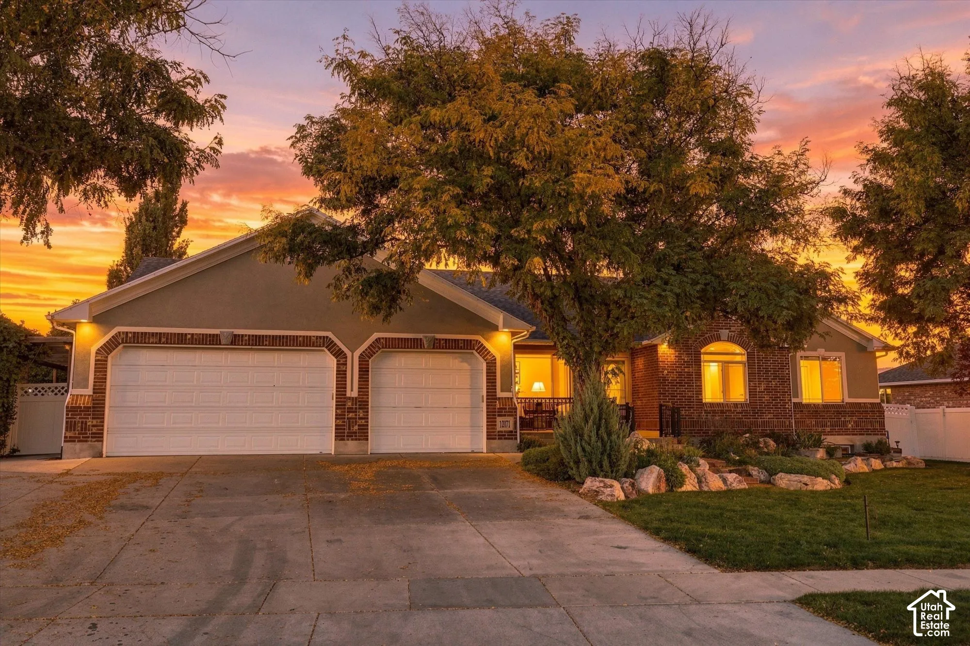 Stunning single-story home featuring a three-car garage with decorative trim detailing, brick and stucco exterior, arched entryway accents, mature landscaping with established trees, and illuminated architectural lighting that highlights the home's elegan