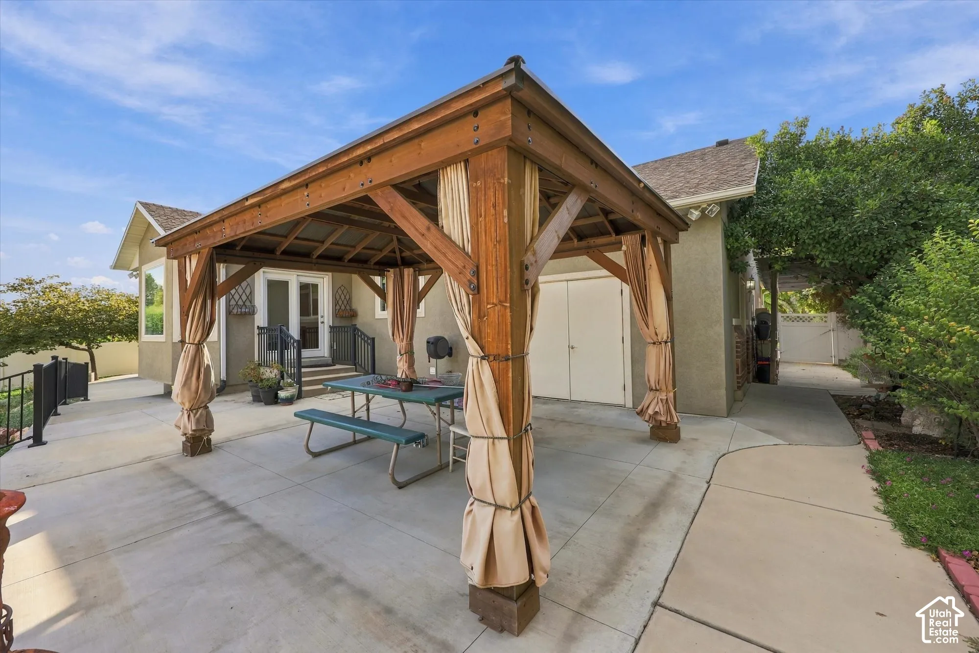 Wood-beam gazebo with privacy drapes, concrete patio, exterior double doors, and a decorative railing