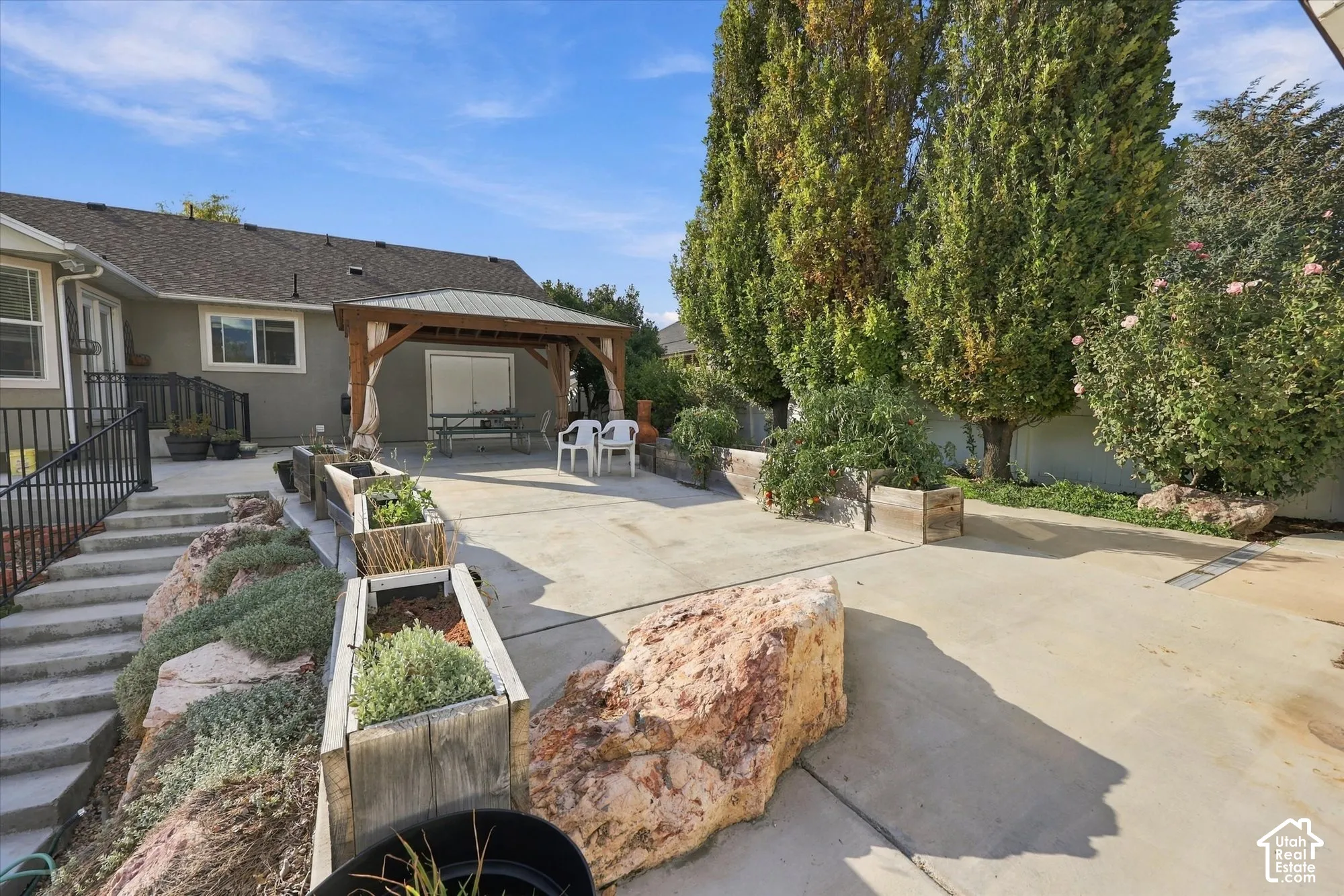 Expansive concrete patio with mature columnar evergreen trees, raised planter beds with drought-tolerant landscaping, and a covered pergola structure