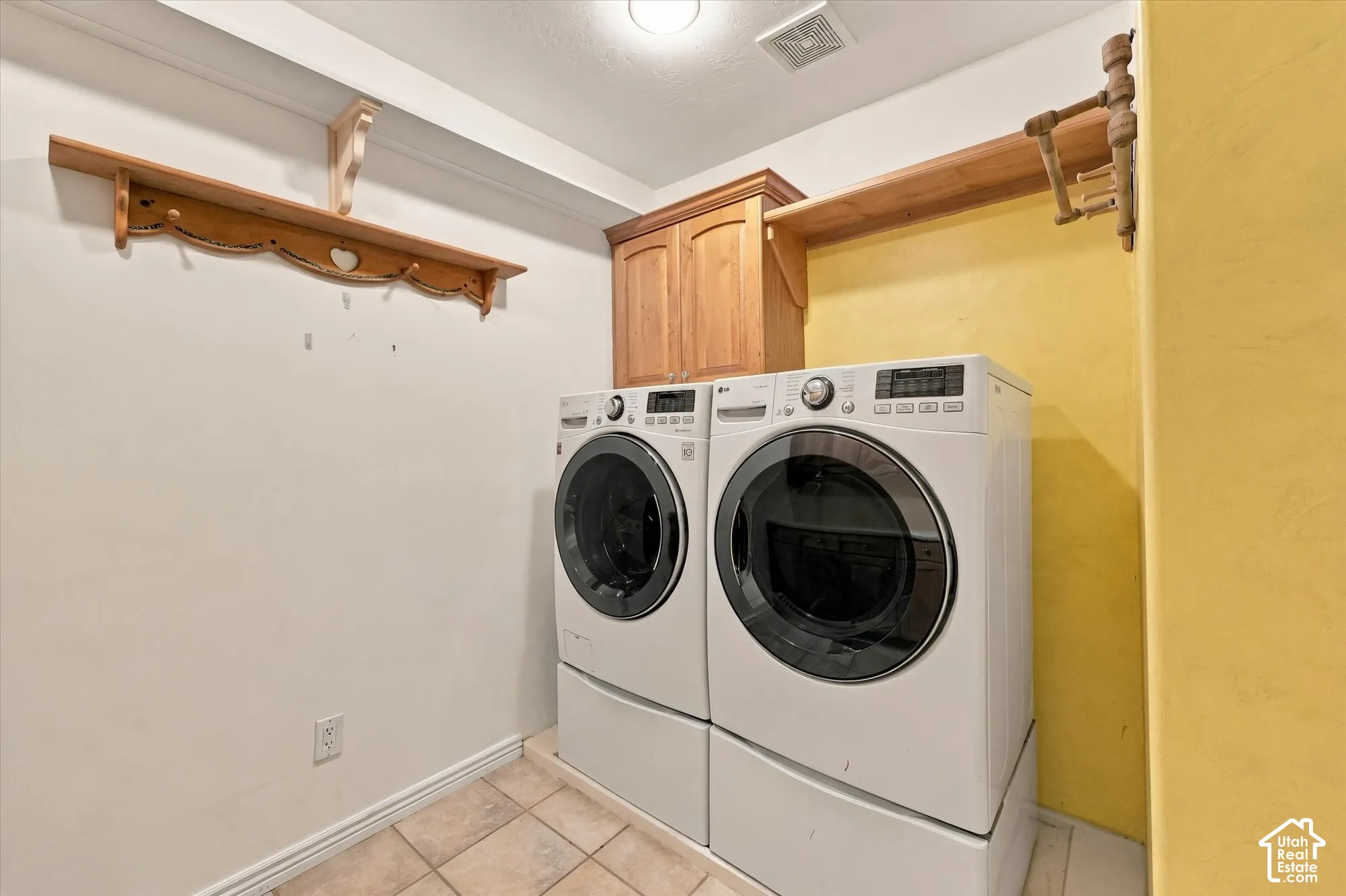 Dedicated laundry room featuring front-load washer and dryer with digital controls, tile flooring, wooden shelving with brackets for storage, and soft yellow accent wall creating a bright, functional utility space