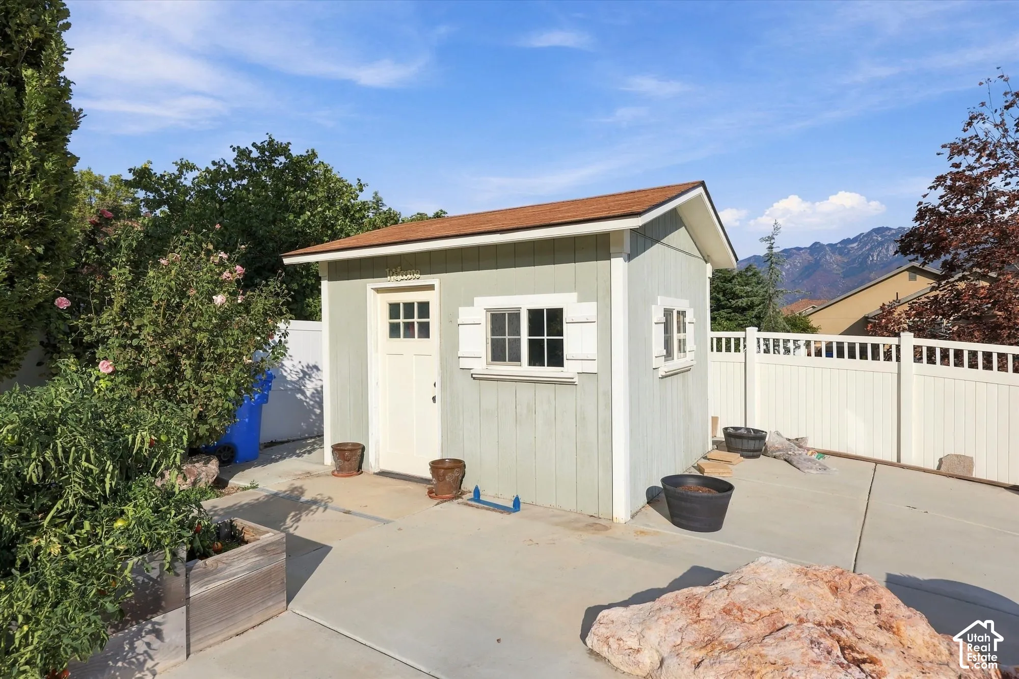Light-colored storage shed with a brown shingle roof, white trim, and a white paneled door