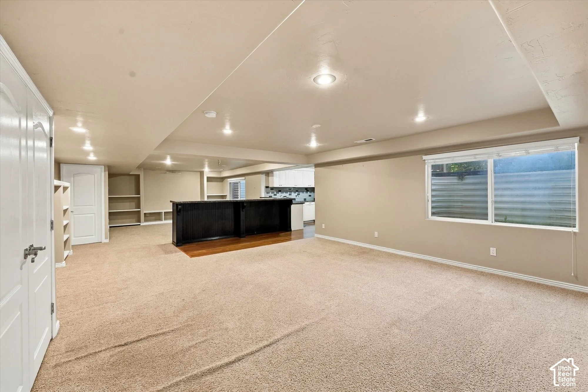 Spacious open-concept living area featuring recessed ceiling lighting, neutral carpet flooring, black kitchen cabinetry with countertop workspace, large horizontal windows with frosted glass panels, and built-in shelving along the perimeter walls