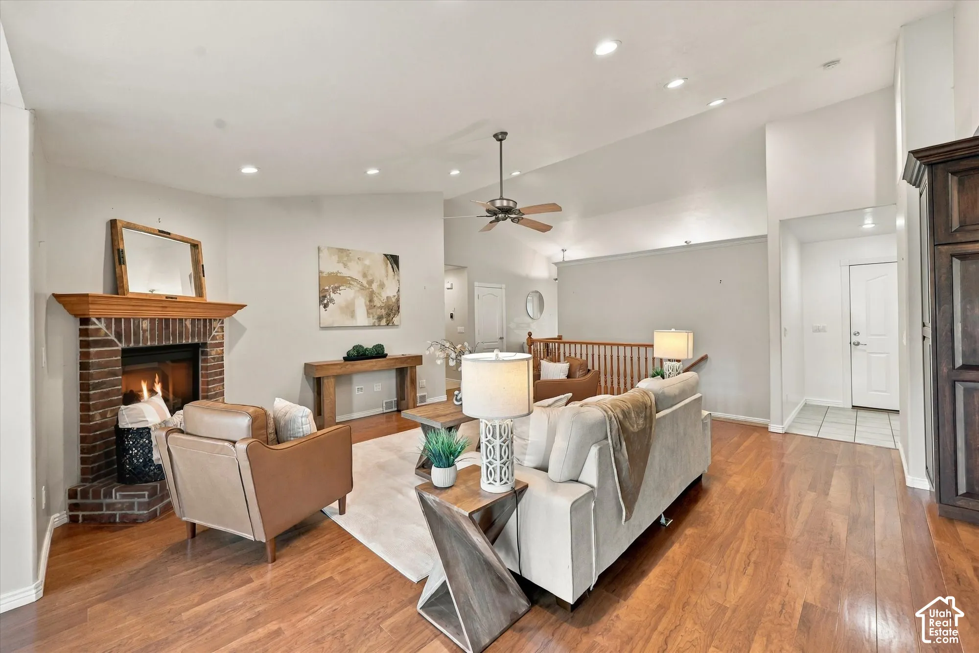 Spacious living room featuring hardwood flooring, a wood-burning fireplace with brick surround, ceiling fan with pendant lighting, and an open floor plan with abundant natural light from recessed ceiling fixtures