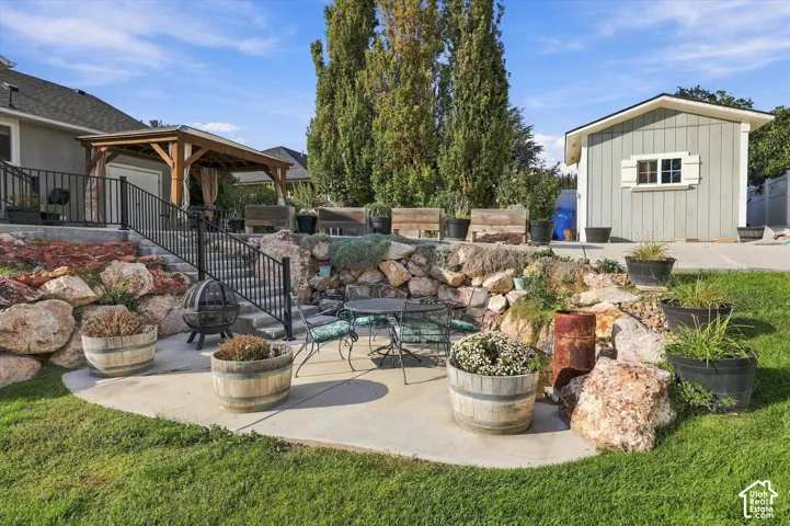 Expansive backyard oasis featuring a concrete patio with integrated seating area, decorative boulder landscaping, mature trees including a tall Italian cypress, multiple large planters with seasonal plantings, wooden pergola structure attached to the home