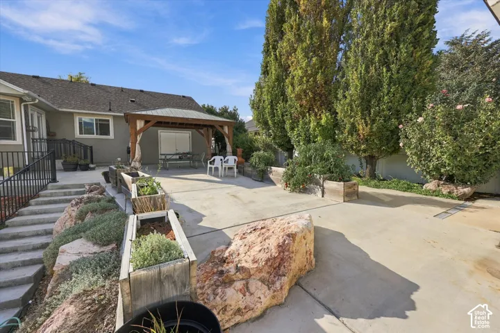 Expansive concrete patio with mature columnar evergreen trees, raised planter beds with drought-tolerant landscaping, and a covered pergola structure