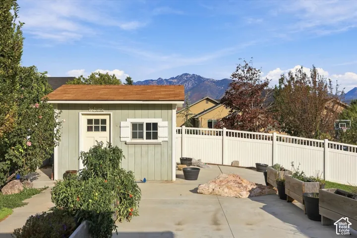 Charming backyard retreat featuring a detached shed with cream-colored vertical siding and gabled roof, white vinyl fencing with decorative balustrade detailing, concrete patio, and mature landscaping with potted plantings and natural boulders