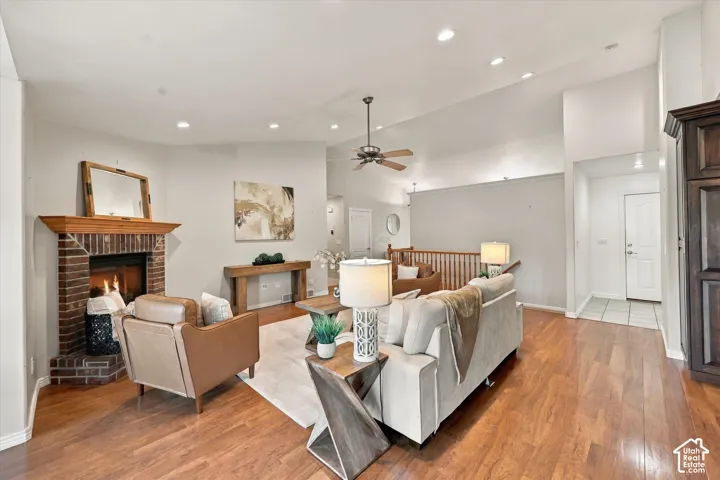 Spacious living room featuring hardwood flooring, a wood-burning fireplace with brick surround, ceiling fan with pendant lighting, and an open floor plan with abundant natural light from recessed ceiling fixtures