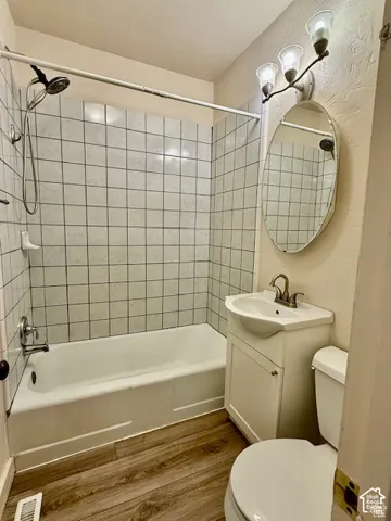 White subway tile shower surround, white bathtub, wood-finish flooring, white vanity with integrated sink, and wall-mounted light fixture