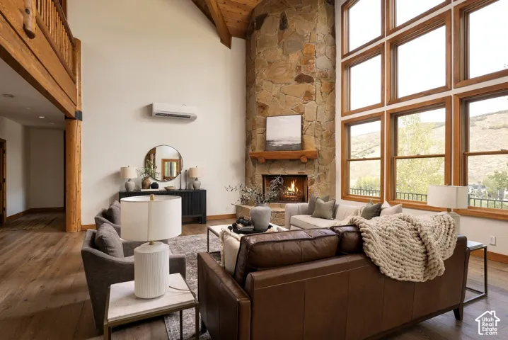 Vaulted ceiling with exposed wood beams, stone fireplace with a wood mantel, large multi-pane windows, and wood-finish flooring