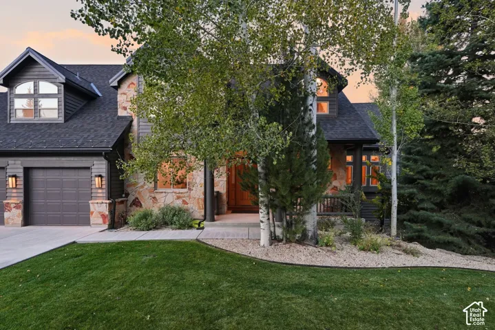 Contemporary home featuring a two-car garage with dark panel doors, modern architectural detailing with mixed exterior materials including brick and dark siding, expansive manicured lawn with mature landscaping, and mature trees providing natural screenin