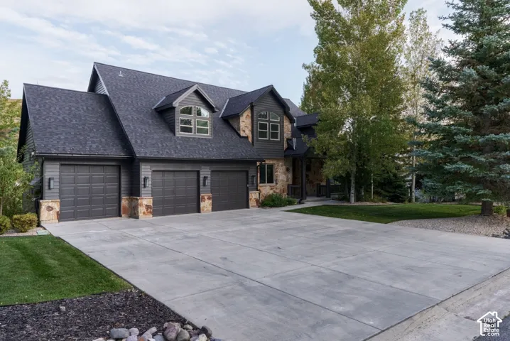 Exterior featuring a multi-car garage, stone accents, dormer windows, a large driveway, and mature landscaping
