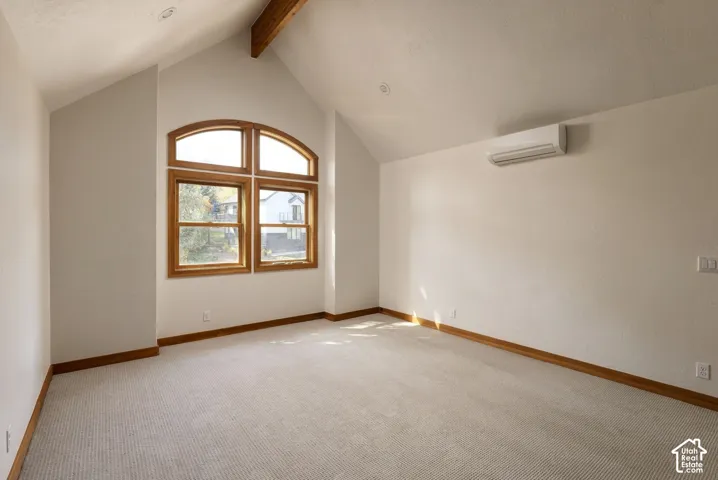 Vaulted ceiling with an exposed wood beam, large arched window with wood framing, wall-mounted climate control unit, textured carpet flooring, and wood baseboards