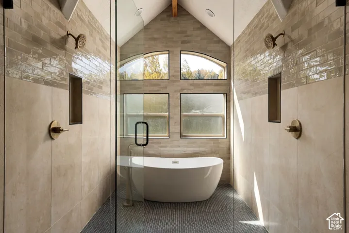 Vaulted ceiling with exposed wood beam, freestanding soaking tub, dual walk-in showers with glass enclosures, mosaic tile shower flooring, and arched window