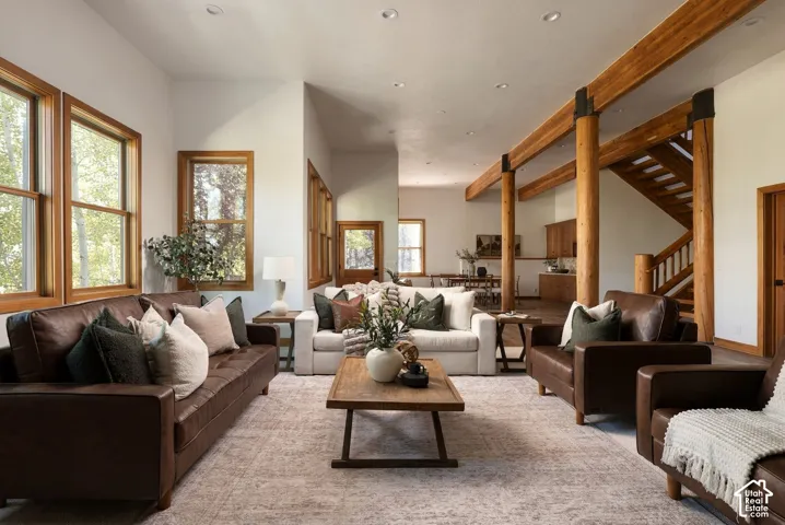 Open-concept living space with exposed wood beams, wood-framed windows, recessed lighting, and a prominent wood staircase