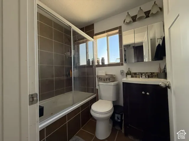 Tub with sliding glass enclosure, brown tile surround, dark wood-finish vanity with white basin, medicine cabinet, and window with brown tile trim