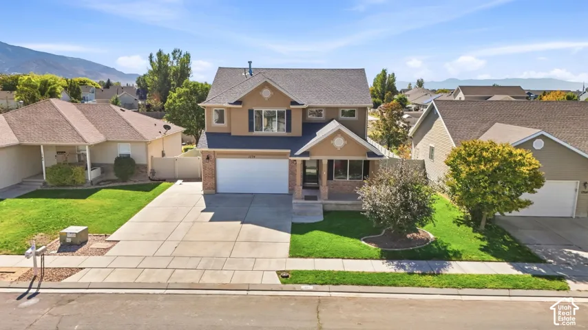 Two-story brick and stone exterior with blue accent siding, featuring a two-car garage, manicured front lawn with mature landscaping, and mountain backdrop