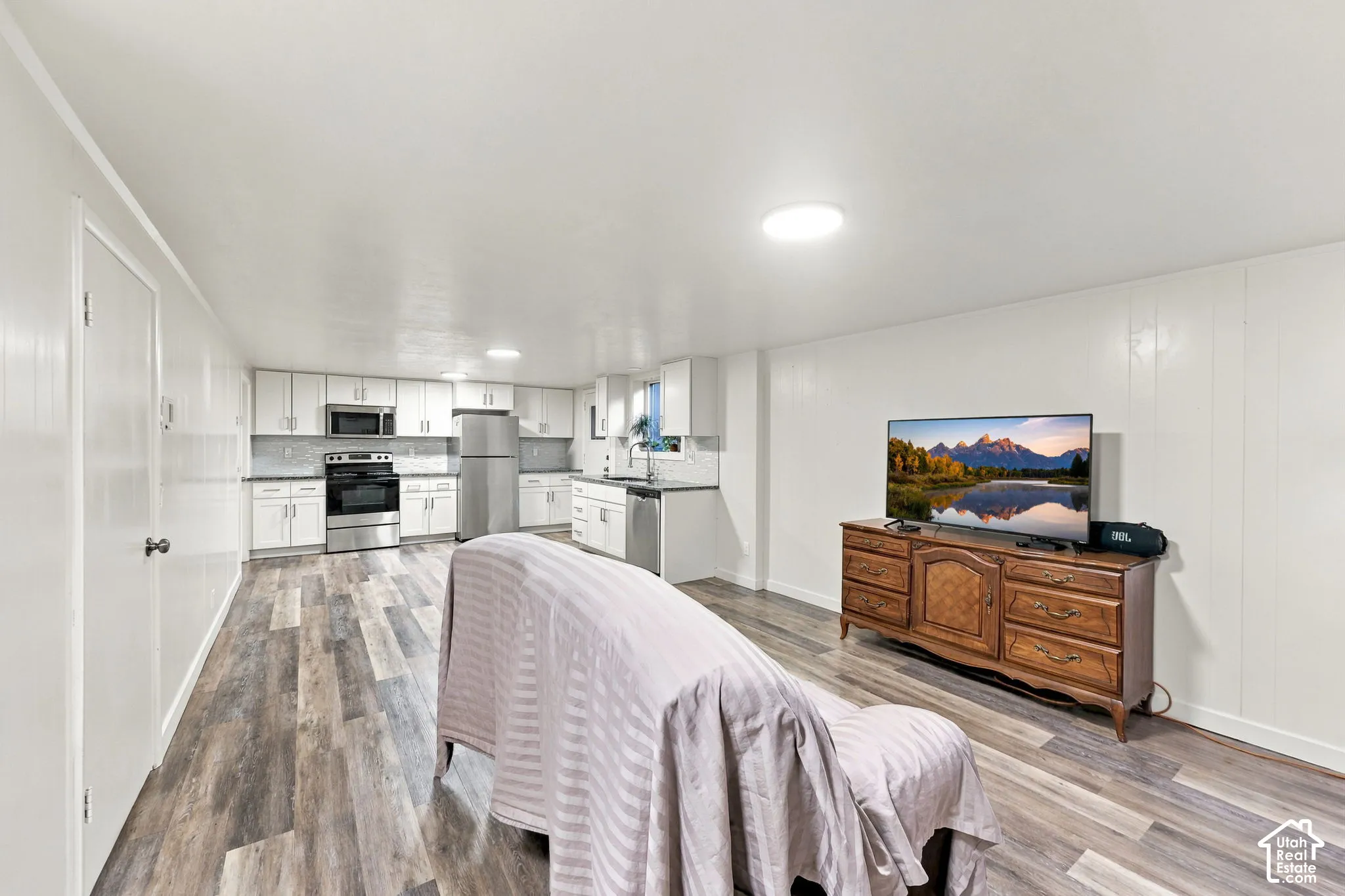 Open-concept living space featuring wood-finish flooring throughout, white cabinetry with stainless steel appliances, recessed lighting, and a wood dresser with mounted television