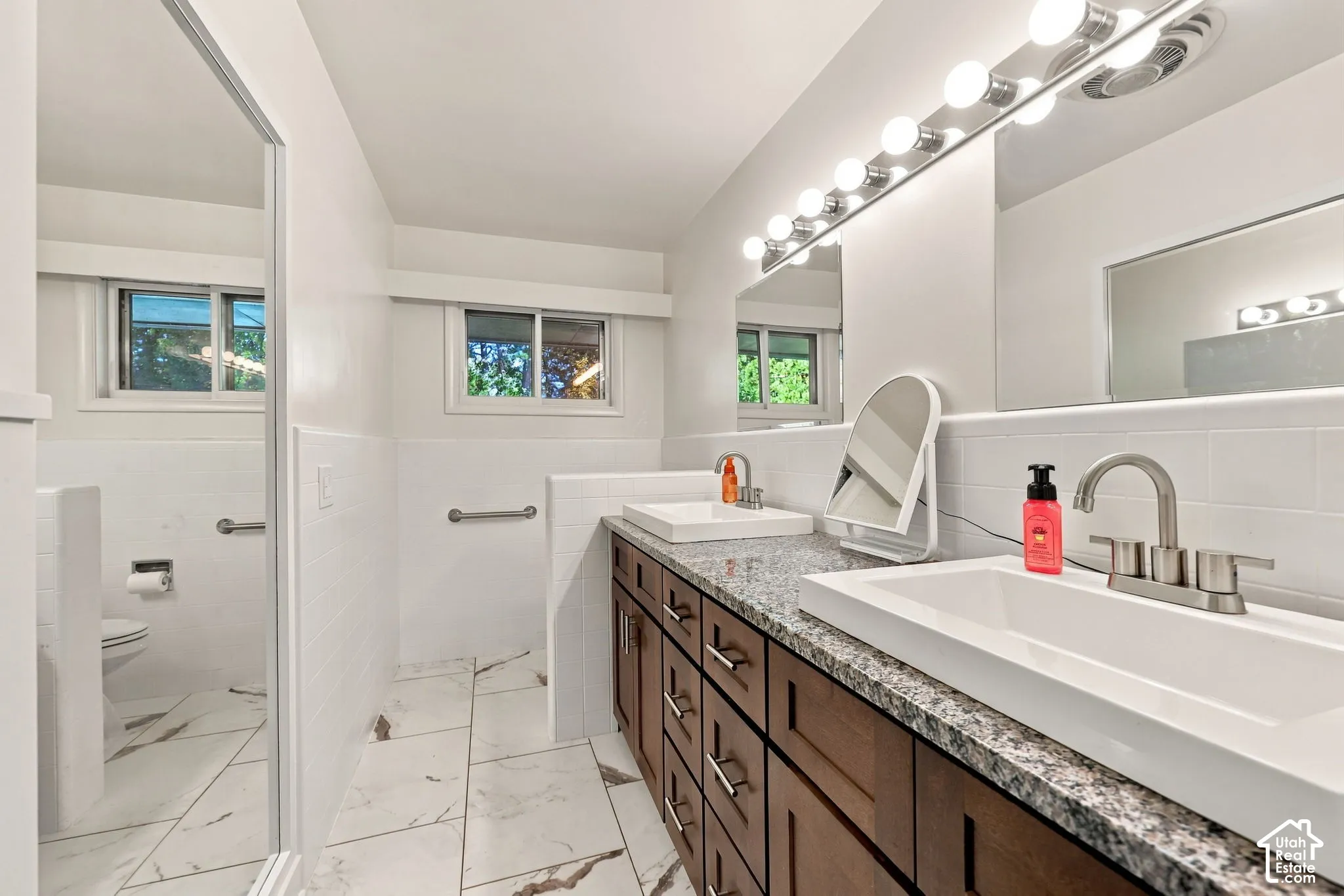 Dual undermount sinks with brushed nickel fixtures, granite-finish countertop, dark wood-finish cabinetry, vanity lighting with multiple bulbs, and large format floor tiling