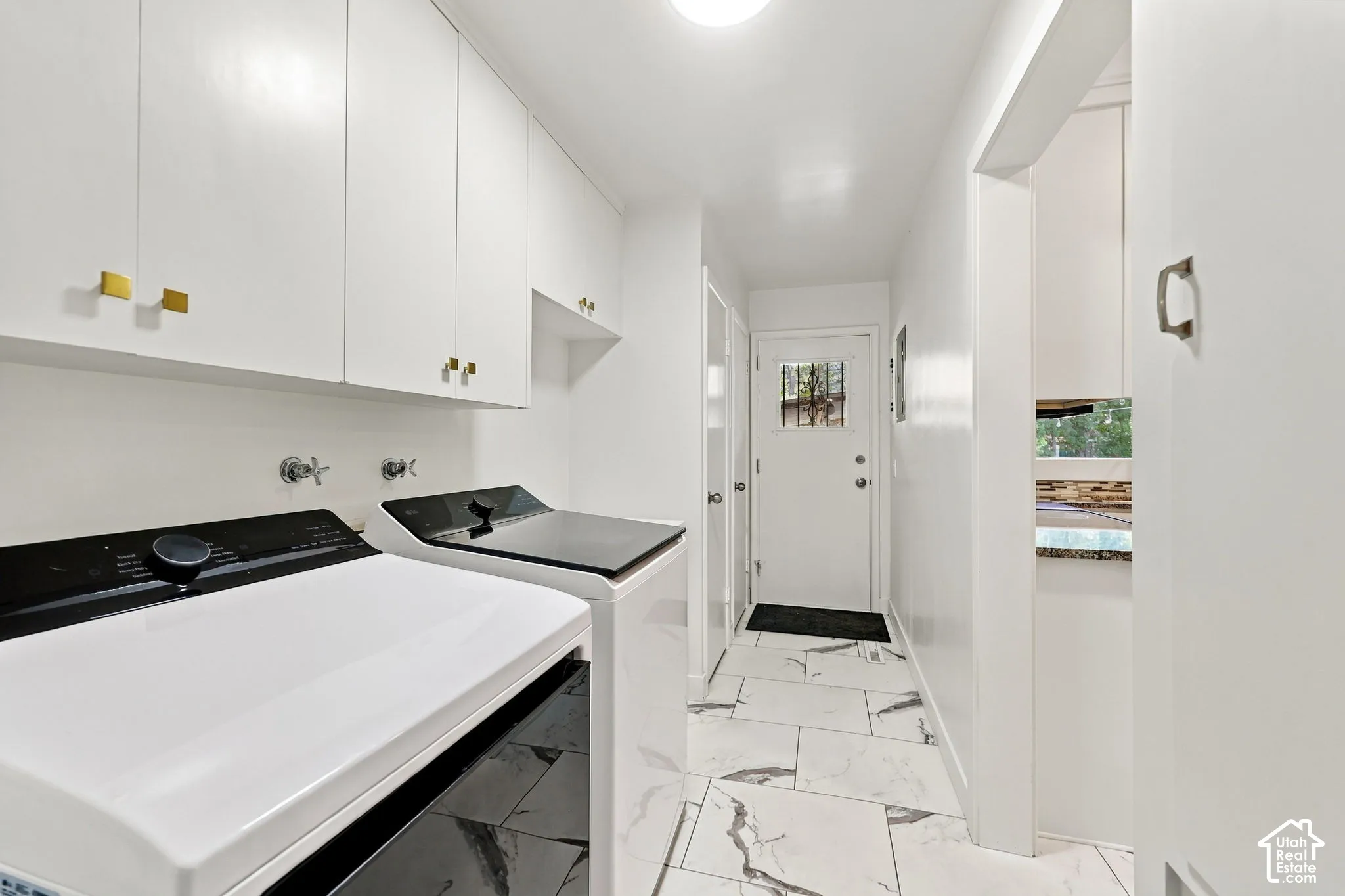 Modern laundry room featuring side-by-side front-load washer and dryer with black and white finishes, white cabinetry with brass hardware, marble-look tile flooring, and recessed ceiling lighting throughout the bright, linear space