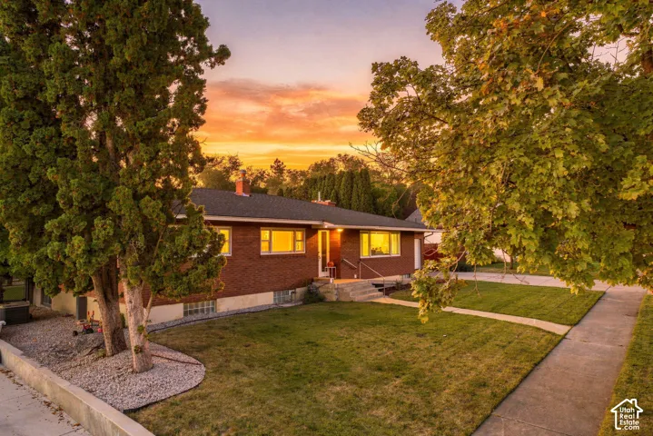 Brick exterior with multiple windows, mature trees, a manicured lawn, and a concrete walkway