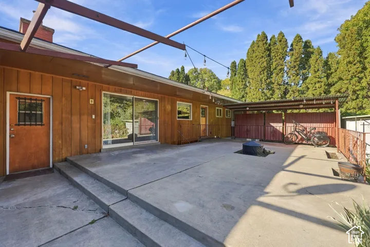 Modern wood-siding residence with expansive concrete patio featuring pergola overhead structure, large multi-pane windows, and mature evergreen trees framing the property