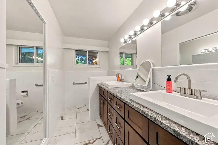 Dual undermount sinks with brushed nickel fixtures, granite-finish countertop, dark wood-finish cabinetry, vanity lighting with multiple bulbs, and large format floor tiling