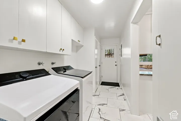 Modern laundry room featuring side-by-side front-load washer and dryer with black and white finishes, white cabinetry with brass hardware, marble-look tile flooring, and recessed ceiling lighting throughout the bright, linear space