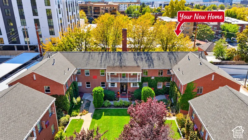 U-shaped brick exterior building featuring a central courtyard, shingled roofing, second-story balcony with decorative metal railing, and mature landscaping