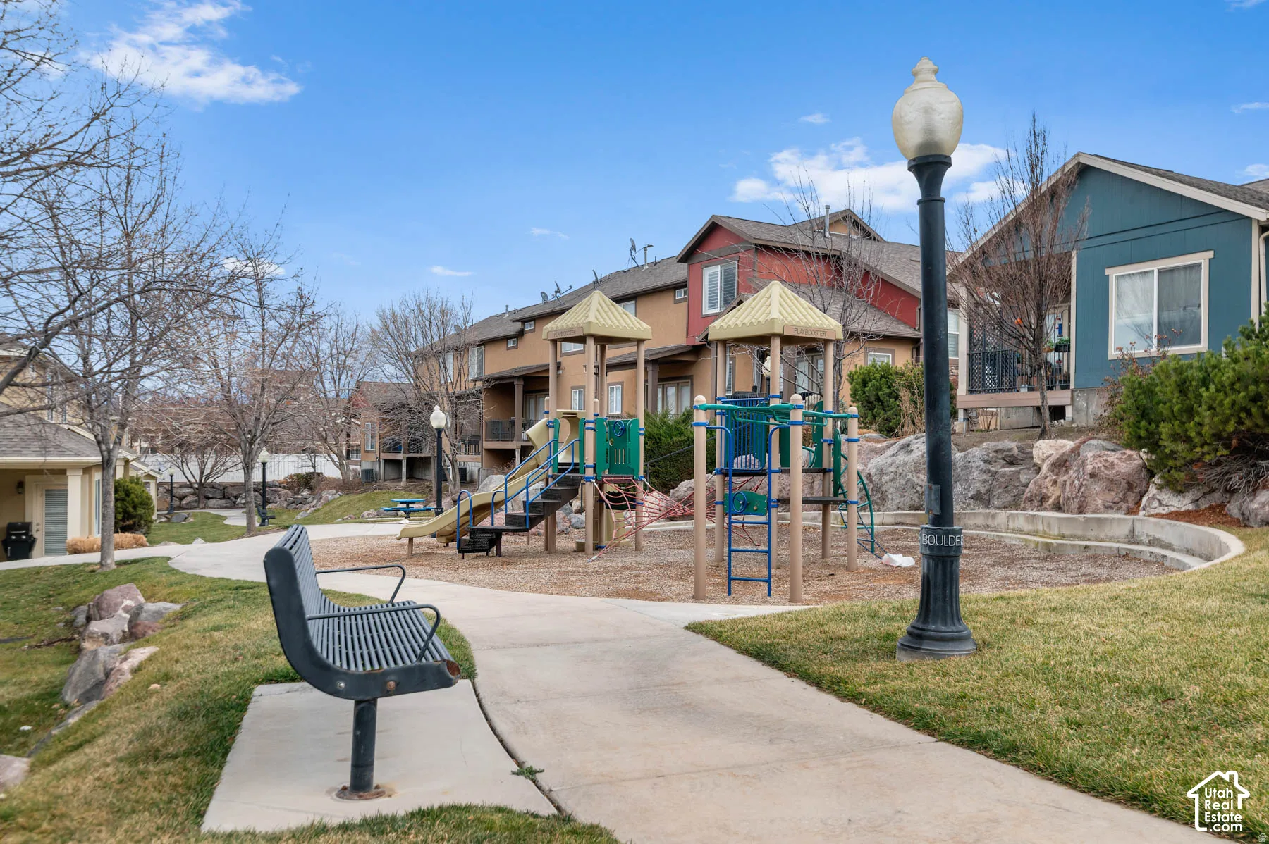 Outdoor recreation area featuring a playground structure, concrete pathways, metal seating, and decorative lamp posts set within a landscaped common area
