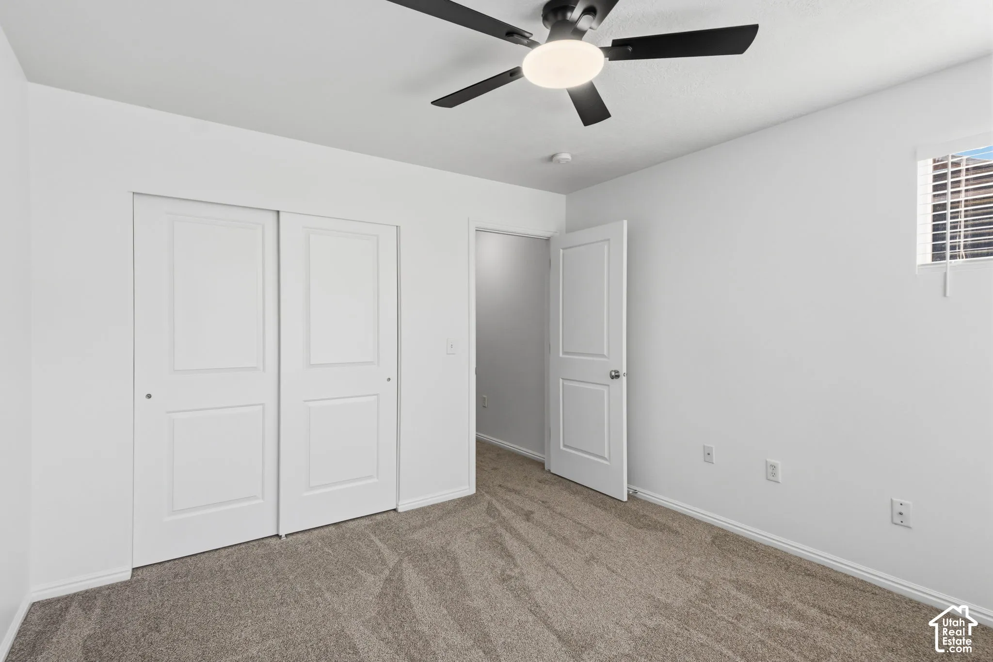 Neutral wall color, ceiling fan with integrated lighting, window with horizontal blinds, textured carpet, and a white paneled door