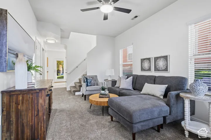 Open-concept living space with textured carpeting, white walls, multiple windows with blinds, a ceiling fan with integrated lighting, and a staircase with a white railing