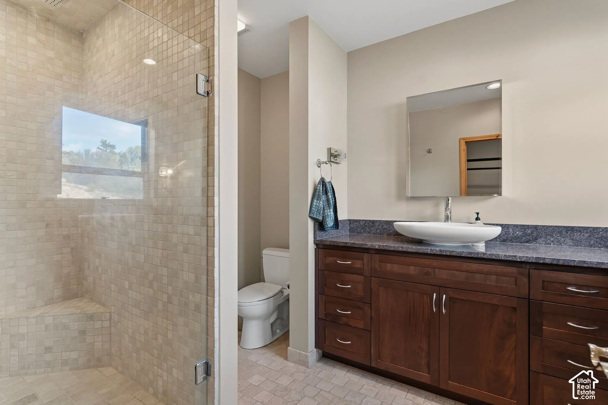 Spa-inspired primary bathroom featuring a frameless glass-enclosed walk-in shower with tile surround, dual-sink vanity with wood cabinetry and dark countertop, large mirror with integrated lighting, and neutral tile flooring throughout