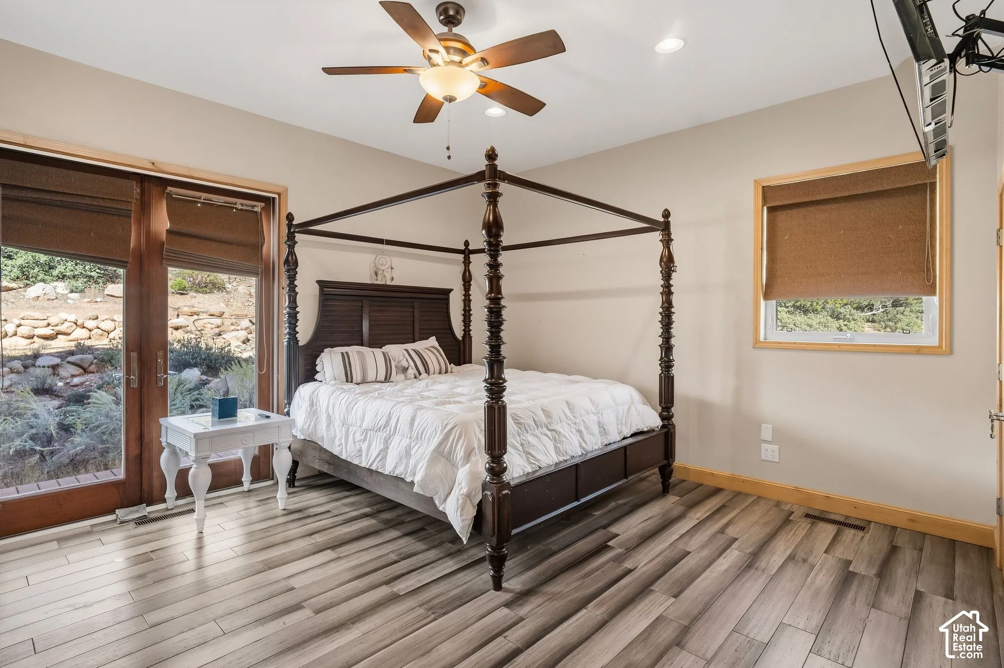 Primary Bedroom Suite featuring a four-poster bed frame, wood-finish flooring with neutral striping, ceiling fan with integrated lighting, and abundant natural light from dual glass doors and transom window