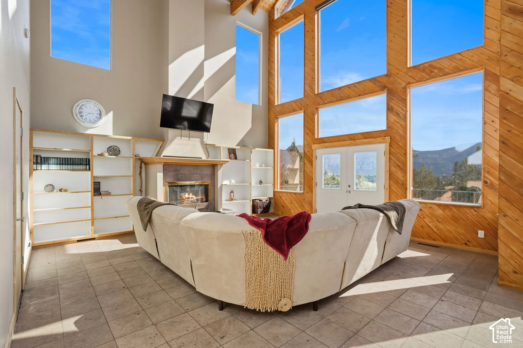 Vaulted ceiling with exposed wood beams, floor-to-ceiling windows, wood-paneled accent wall, built-in shelving, and a tiled fireplace