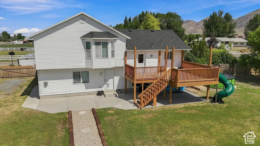 Two-story residence with white vinyl siding and gabled roofline, featuring a multi-level wooden deck with stairs, mature landscaping including evergreen and deciduous trees, expansive lawn, and detached outbuildings