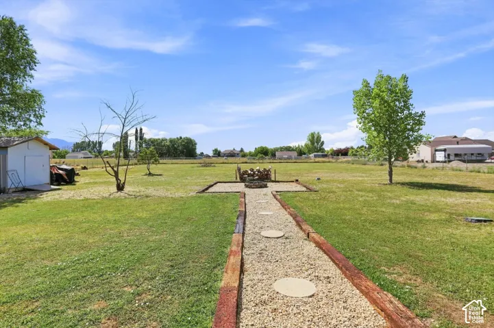 Expansive grassy yard featuring a gravel pathway with circular stepping stones, a built-in fire pit, mature trees, and a storage shed