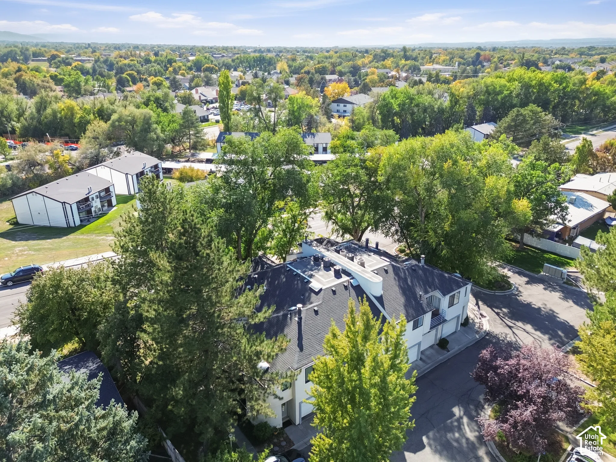 Aerial perspective showcasing a multi-unit property with a dark shingle roof, surrounded by mature trees, and an open sky