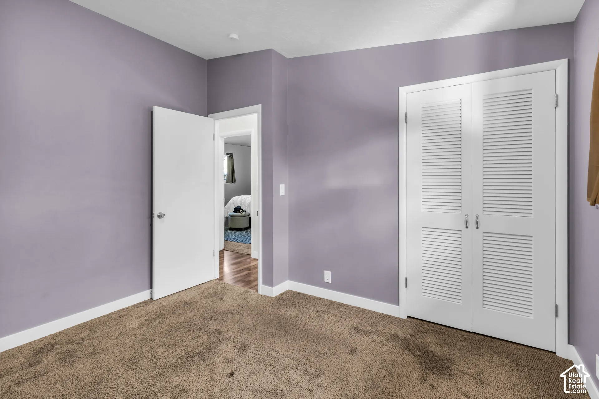 Plush brown carpet flooring, light purple wall paint, white trim, white door with brushed nickel hardware, and white louvered bifold doors