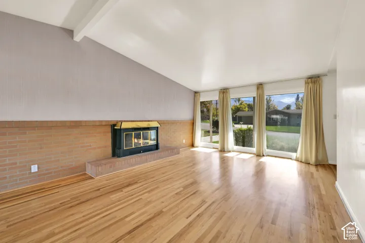 Vaulted ceiling with exposed beam, brick fireplace with a raised hearth, wood-finish flooring, and a wall of windows