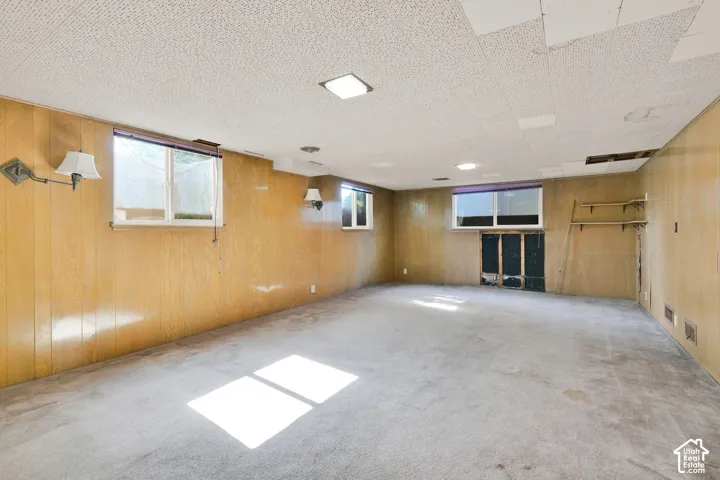 Spacious basement recreation room featuring wood-paneled walls, recessed and wall-mounted lighting, concrete flooring, and multiple windows providing natural light