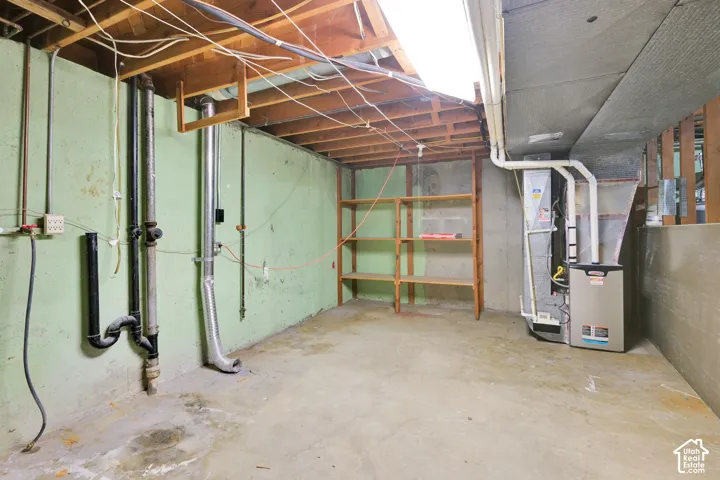 Unfinished basement space featuring exposed ceiling joists, concrete flooring, a utility wall with various pipes, and a furnace unit
