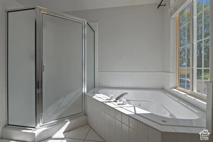 Spa-inspired primary bathroom featuring a corner soaking tub with curved edges, frameless glass shower enclosure with chrome hardware, ceramic tile wainscoting, and abundant natural light from large windows overlooking the grounds