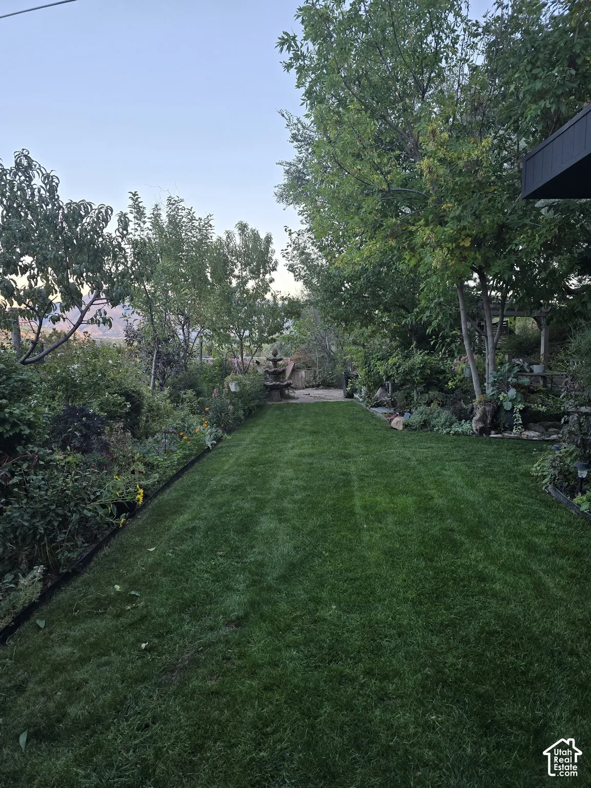 Expansive backyard featuring a manicured lawn with mature shade trees, established hedges and perimeter landscaping, and a covered structure visible at the property's rear