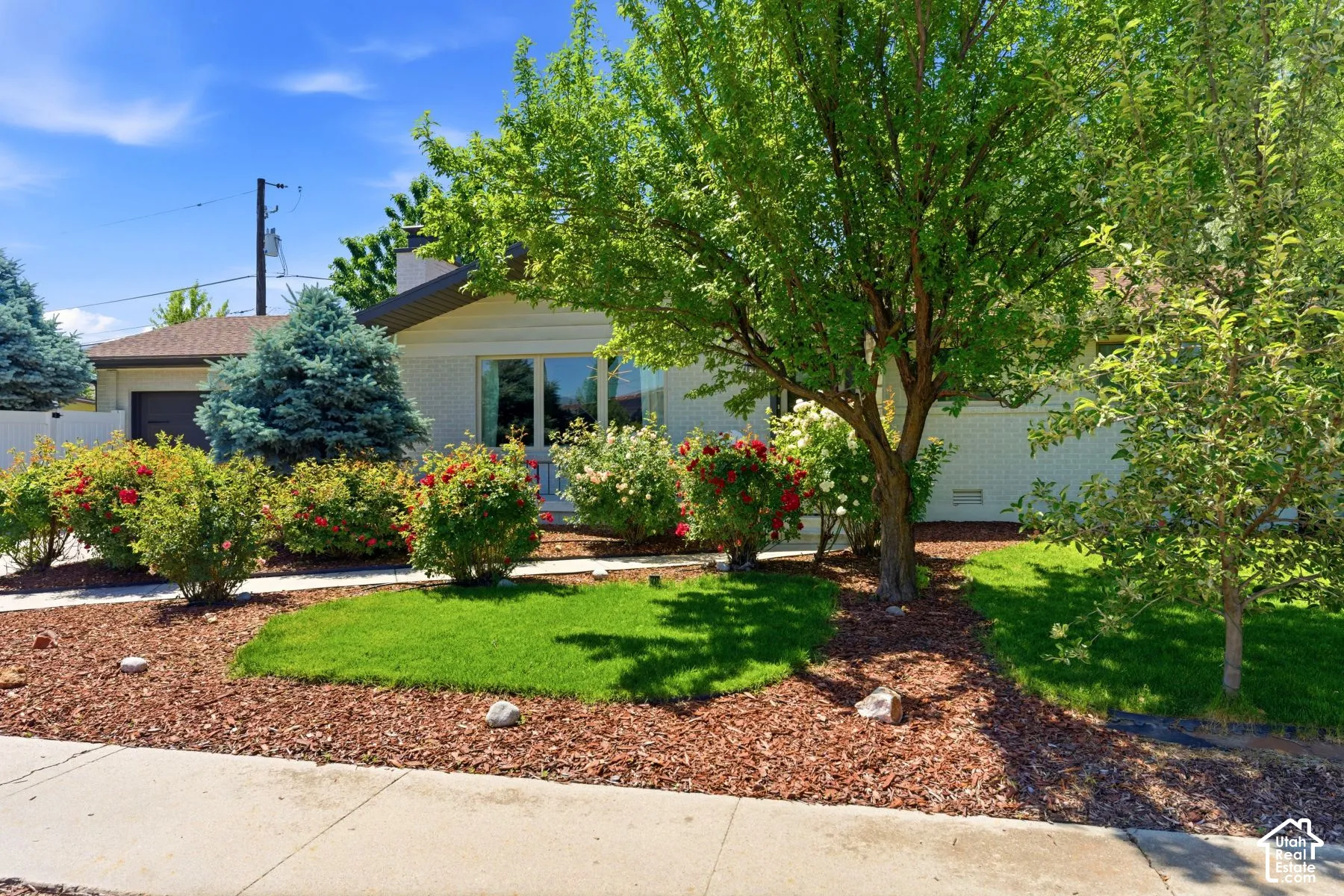 White brick exterior with a dark roofline, mature trees, manicured lawn, and planted shrubbery