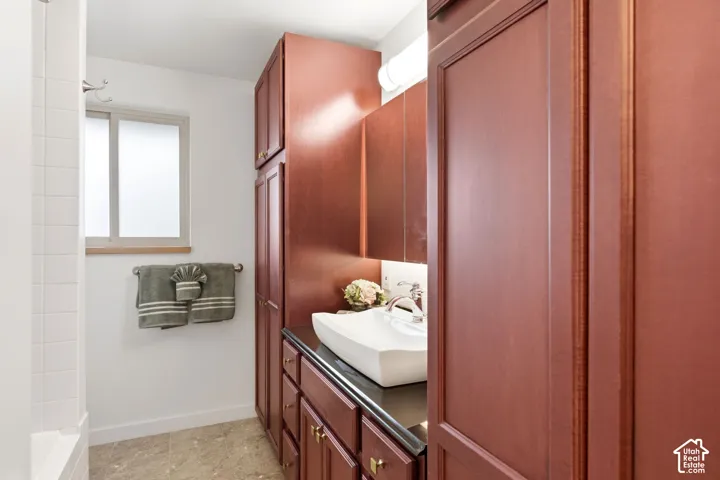 Rich wood-finish cabinetry with golden hardware, a vessel sink, a dark countertop, and tiled flooring