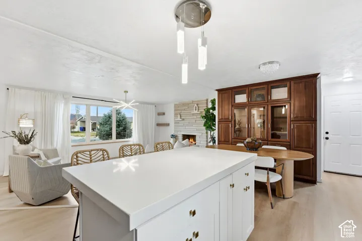 White kitchen island with a solid surface countertop, light wood-finish flooring, built-in display cabinetry, stone-clad fireplace, and large picture window